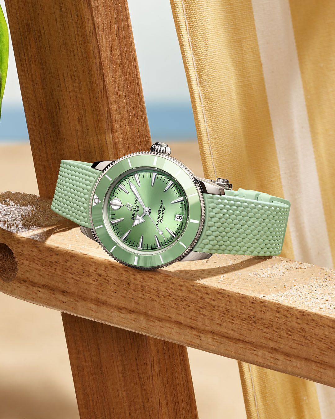 A light green watch sitting on a boardwalk railing in front of a beach