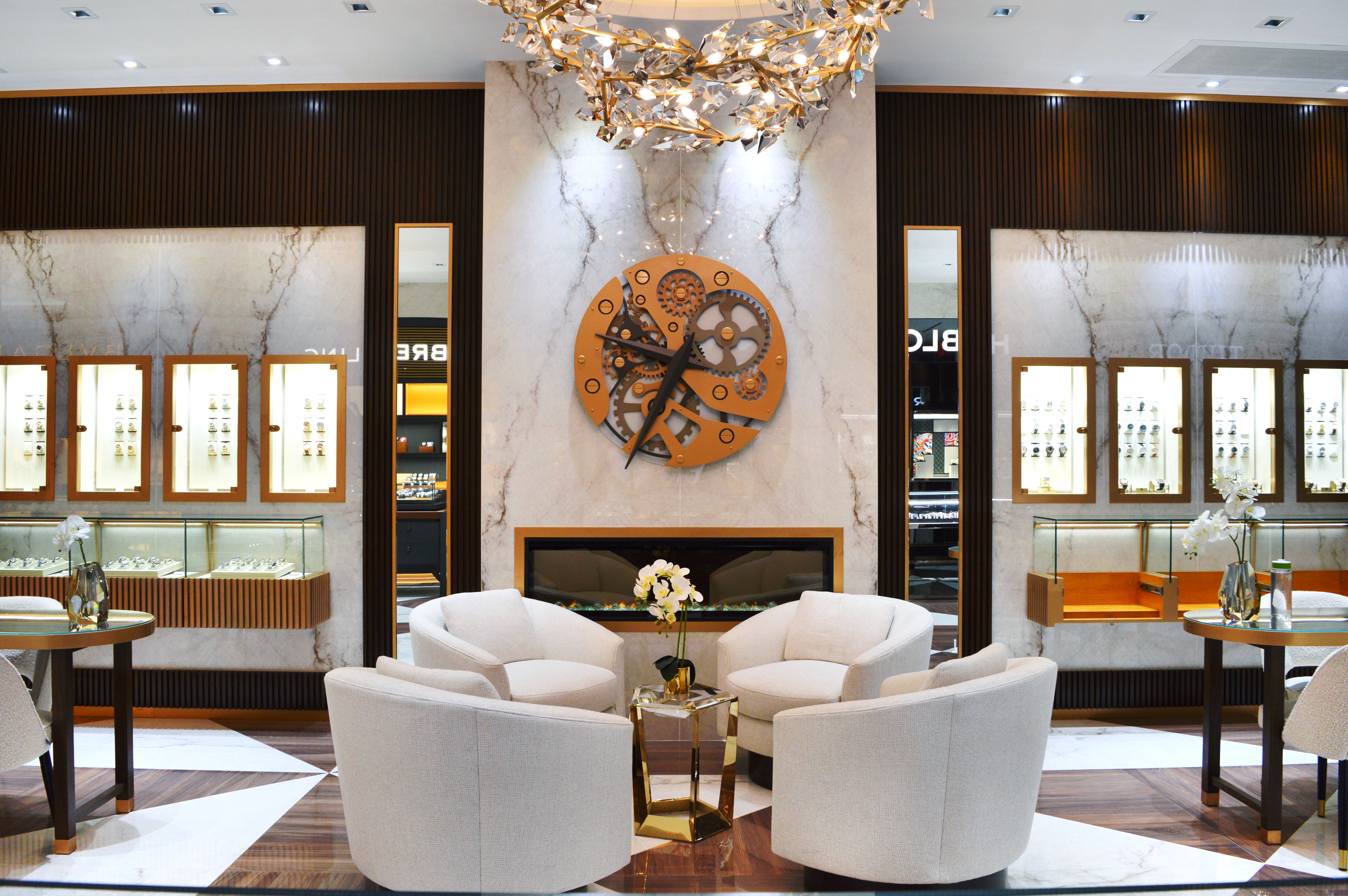 A watch salon with comfy white sofa chairs and a fireplace with an elaborate brass clock hanging over it