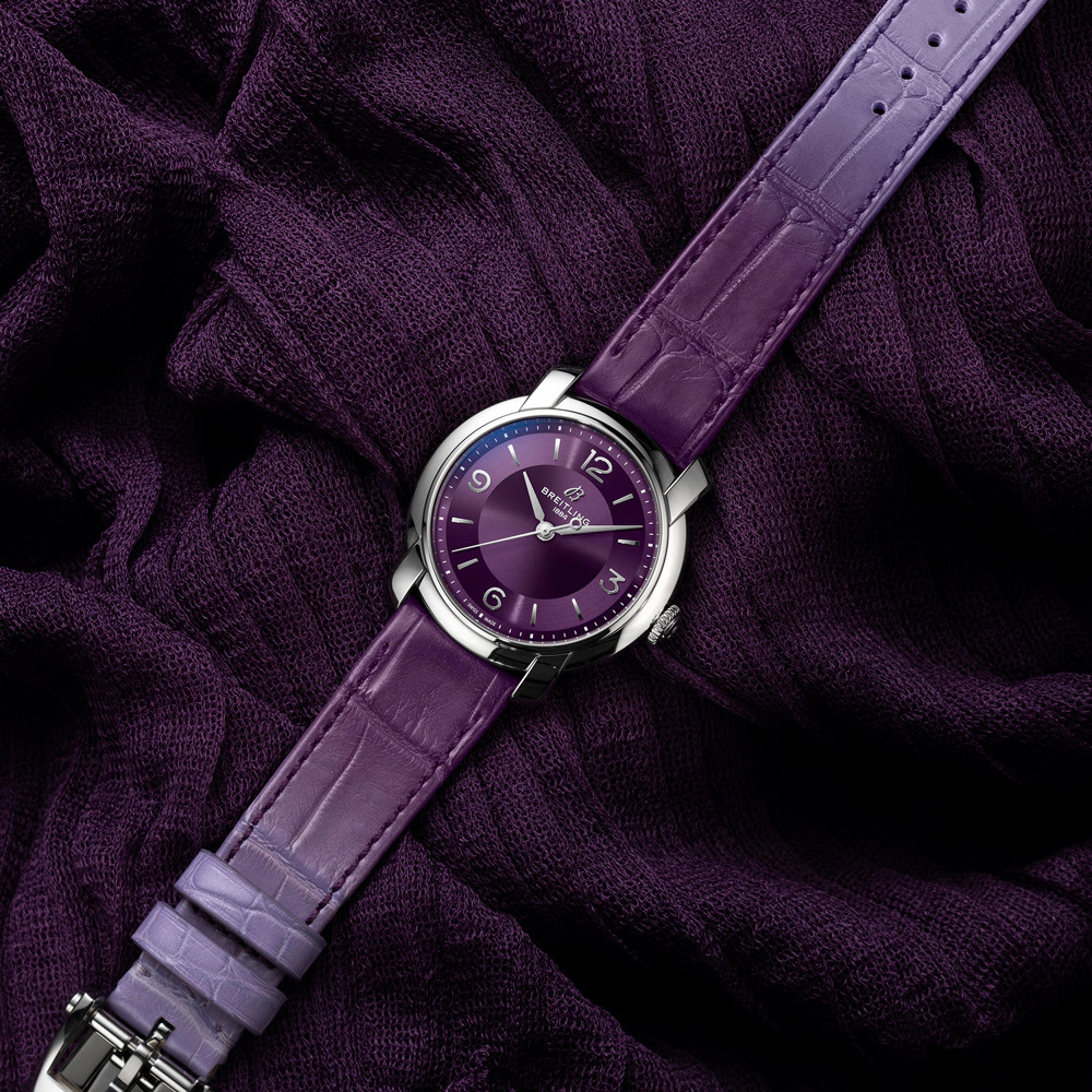 A purple watch with a silver case laying on a textured purple fabric