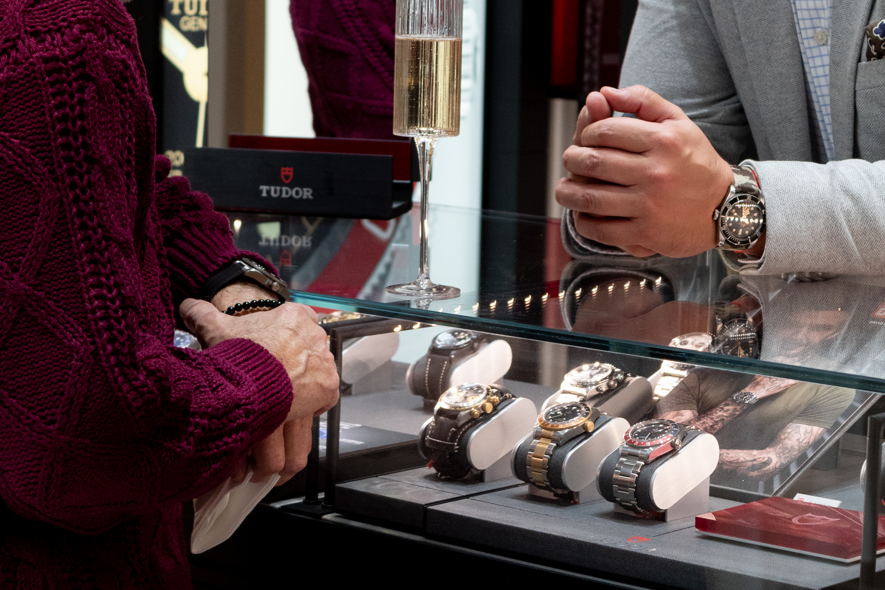 A man in a grey suit assists another man in a burgundy sweater with examining Tudor watches in a case